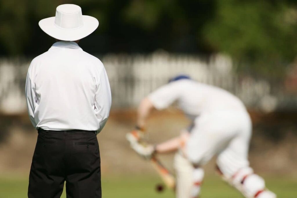 Back side angle of a leg umpire on field with a batsman playing in the background, the background is blurred.