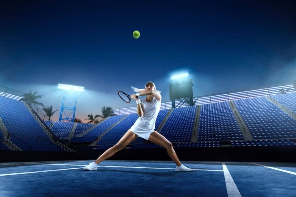 A female tennis player in a white outfit is captured mid-action on a blue hard court at night. She is positioned in a wide stance, preparing to hit a ball that is airborne above her. The stadium is empty with rows of blue seats.