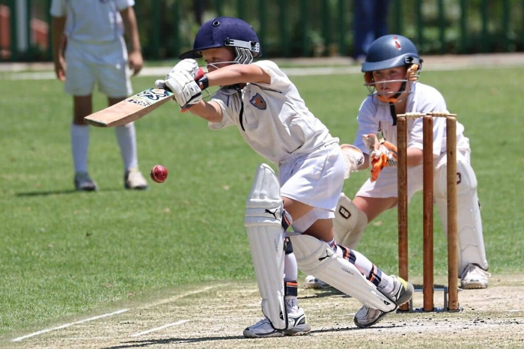Cricket action shot with a batsman in full gear striking the ball mid-air, as the wicketkeeper and fielder stand alert on a grassy pitch.