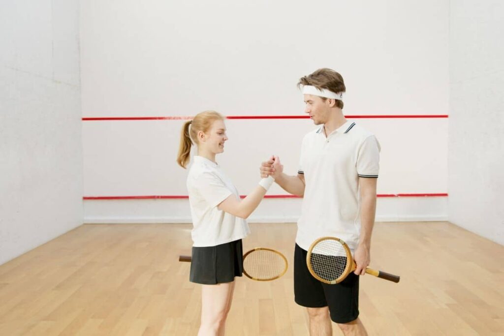 A woman and a man on a squash court giving each other a friendly handshake, both smiling and dressed in white sportswear. Each is holding a wooden squash racket, and red boundary lines are visible on the court walls in the background.