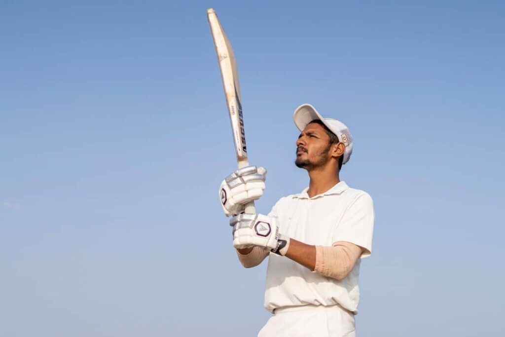 Cricket player in full white kit raising their bat against a clear blue sky, poised for an aerial shot with protective gear and focused form.