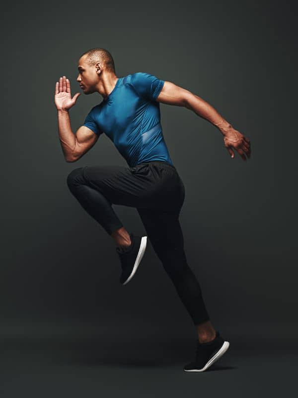 A person in a dynamic running pose, wearing a fitted blue shirt, black athletic pants, and black running shoes with white soles, set against a solid dark background that emphasizes motion and athleticism.