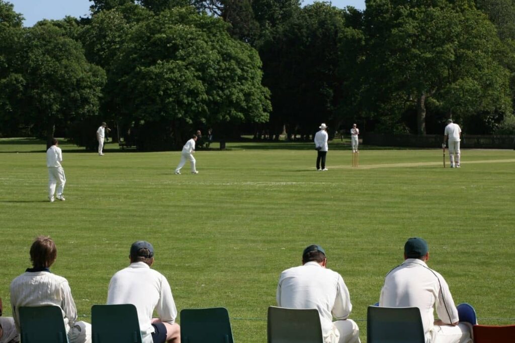 Side view of a cricket match going on. Shot is taken from behind some players sitting, there are trees in the distance.