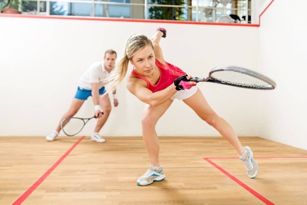 Two players are engaged in a squash match on an indoor court. The player in the foreground, dressed in a pink tank top and white shorts, is lunging forward to strike the ball with a squash racket. Behind them, another player in a white shirt and blue shorts stands ready to respond. The court features a wooden floor, white walls, and red boundary lines.