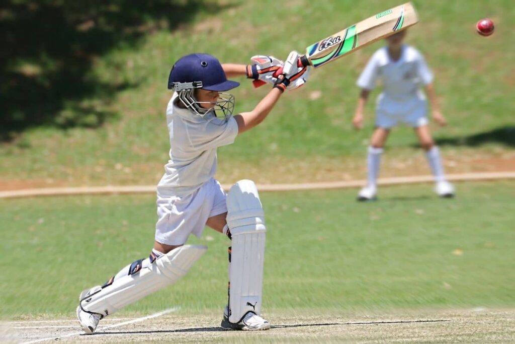 A young cricketer in full white gear and a blue helmet executes a focused shot with a wooden bat on a sunny grass field. The red cricket ball is mid-air, suggesting active play. Another player stands in the background, also dressed in cricket whites, observing or preparing for fielding. The bright setting and body language evoke intensity, technique, and the thrill of a live match.