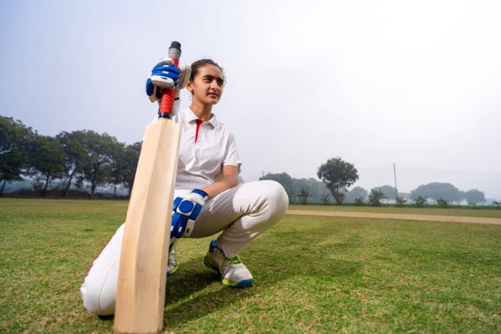 Cricketer kneeling on a grassy pitch in full white kit, gripping a bat upright with both hands in a moment of calm focus before play.