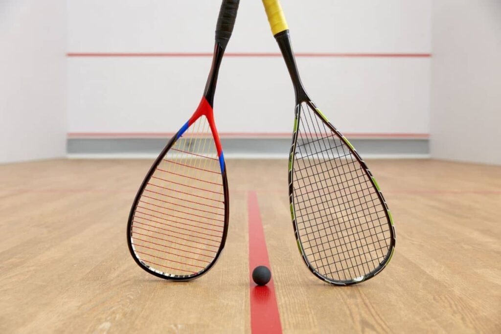 Two squash rackets standing upright with their handles touching and frames angled outward, positioned on a squash court with a black squash ball placed between them on the red center line. Red boundary lines are visible on the white court walls in the background.