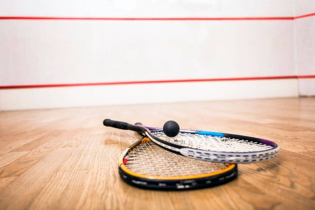 Two squash rackets and a black squash ball resting on a wooden squash court floor, with red boundary lines visible on the white walls in the background.